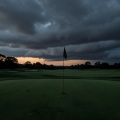 Empty golf course green with a plain flagstick under gathering storm clouds at dusk